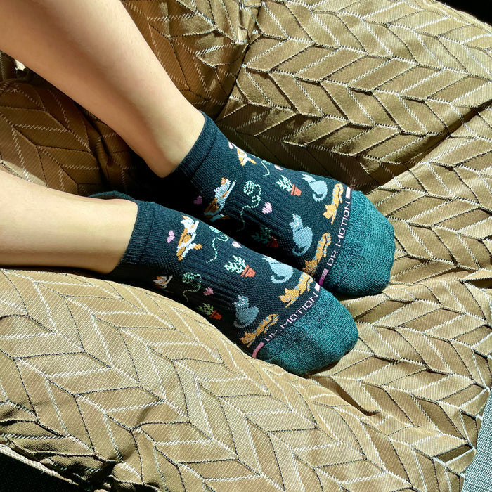 Ankle compression socks with a cat and plant pattern, worn on feet resting on a textured cushion.