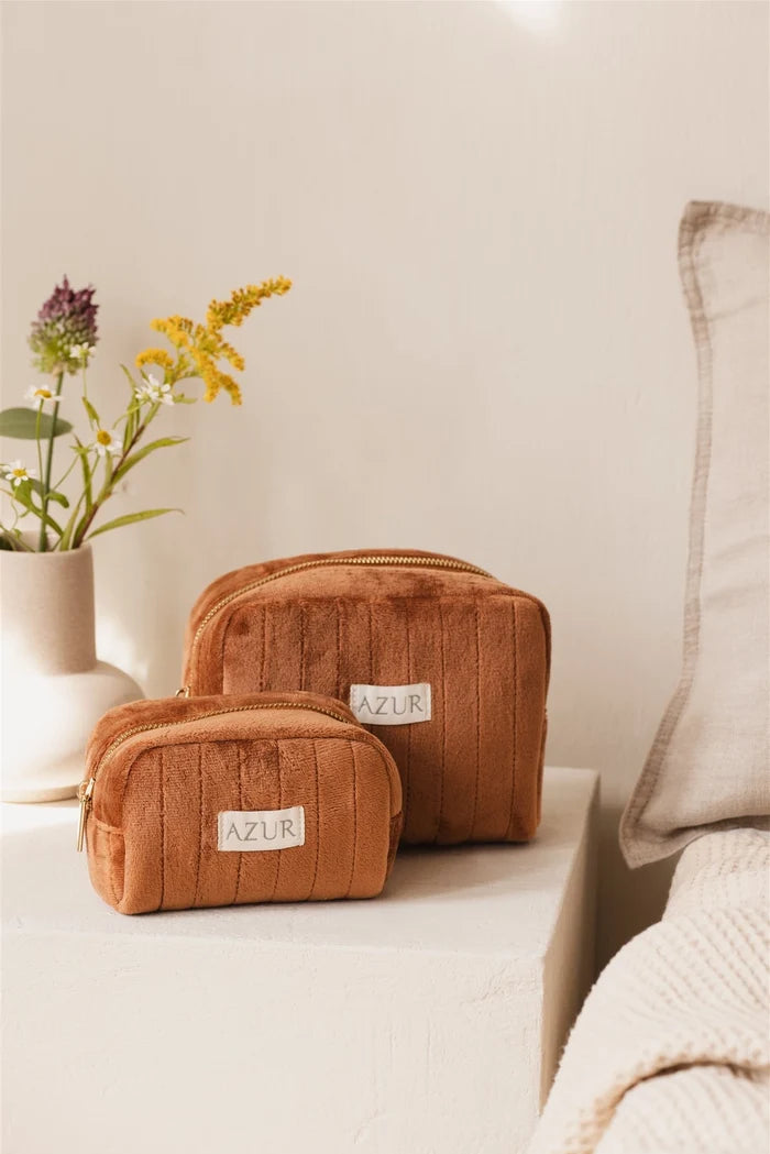Two brown cosmetic bags with 'AZUR' label on a light surface, next to a vase with flowers and a pillow.