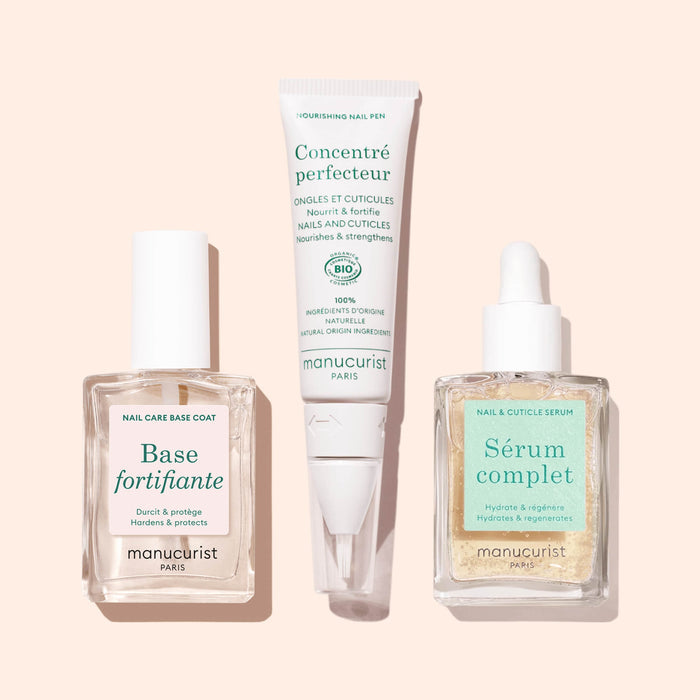 Three Manucurist products on a light pink background: Force Nail Hardener in a square bottle, a nail and cuticle serum with a dropper, and a nourishing nail pen in a tube.