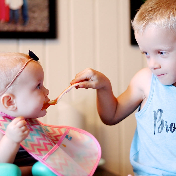 A young boy in a blue tank top carefully feeds a baby girl wearing a headband and colorful bib with the Nuby Hot Safe Baby Spoon (4 Pack), featuring a soft bite tip. The baby sits in a high chair indoors, with both children present.