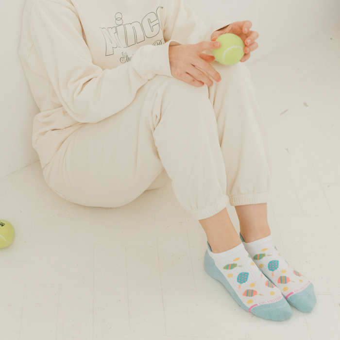 A person sits on the floor wearing cream-colored loungewear and colorful ankle compression socks, holding a tennis ball.