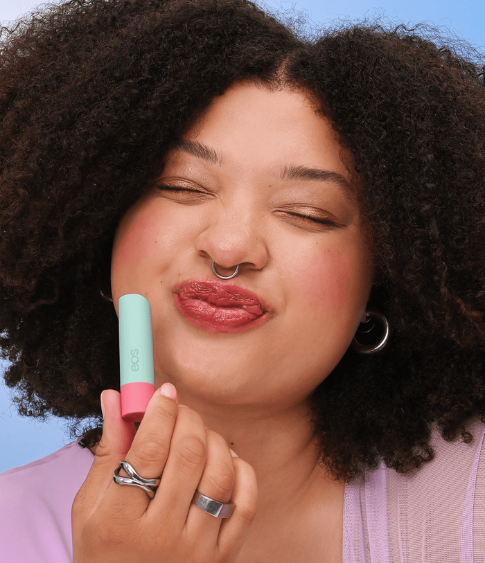 A close-up of a person with curly dark hair applying a pastel mint green and pink lipstick tube to their lips, set against a solid blue background.