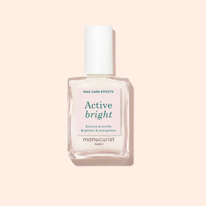 A clear glass bottle of Manucurist Active™ Bright nail polish, featuring a white cap and set against a light beige background. The label highlights that this bio-based nail care product brightens and strengthens nails.