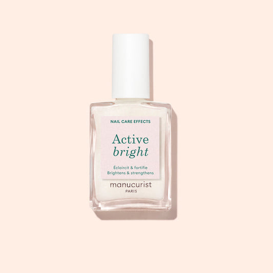 A clear glass bottle of Manucurist Active™ Bright nail polish, featuring a white cap and set against a light beige background. The label highlights that this bio-based nail care product brightens and strengthens nails.