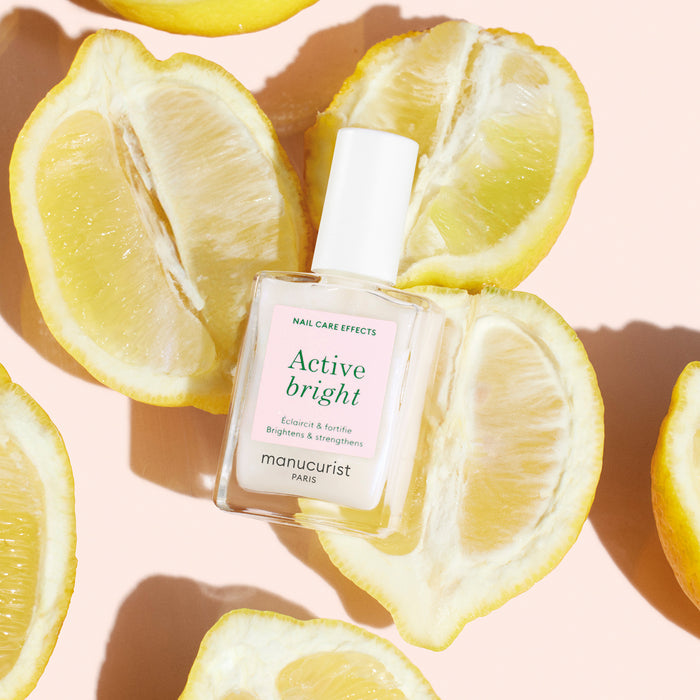 A bottle of Manucurist Active™ Bright nail polish rests on a peach background, encircled by halved lemons arranged in a loose circle. Sunlight filters through, casting gentle shadows from both the lemons and the bottle.