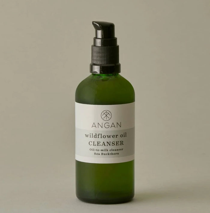A green glass pump bottle of Angan Wildflower Oil Cleanser with a black pump dispenser and a white label, set against a solid neutral gray background.