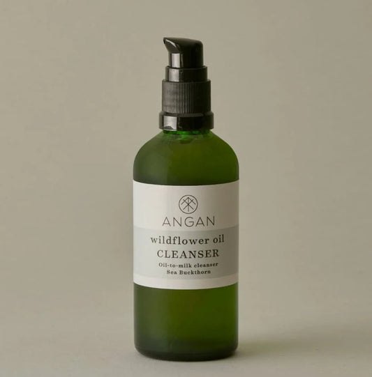 A green glass pump bottle of Angan Wildflower Oil Cleanser with a black pump dispenser and a white label, set against a solid neutral gray background.