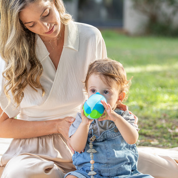 A woman sits outdoors on the grass with a child in denim overalls, who drinks from the Nuby US Baby's First All Silicone Sippy Cup. The green, blurred background highlights their time together.