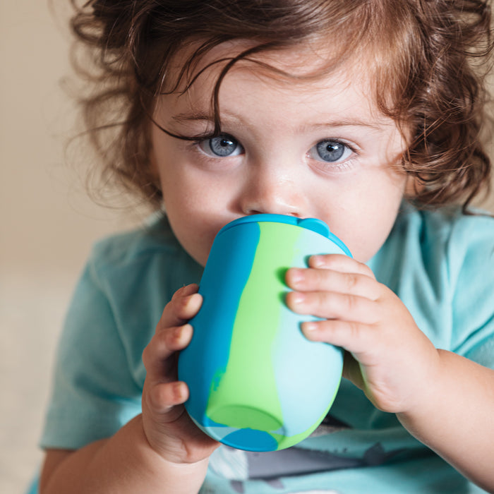 A young child with curly hair and blue eyes drinks from the Nuby US Baby's First All Silicone Sippy Cup, which is blue and green with a free-flow spout, while wearing a light blue shirt.