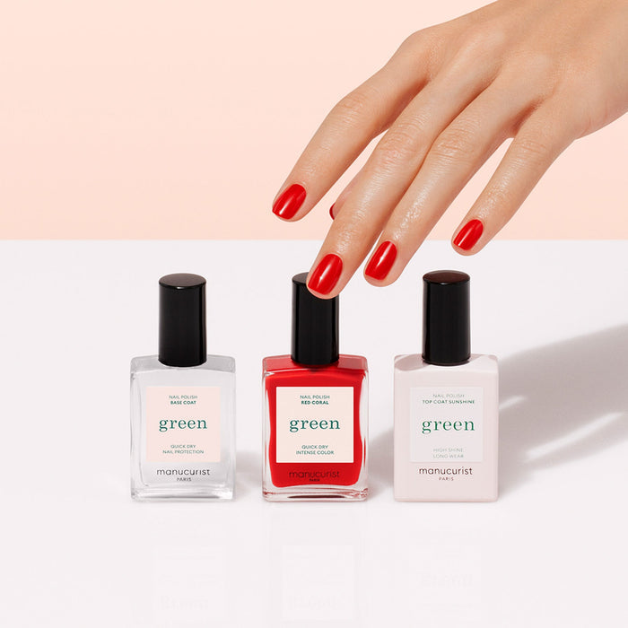 A hand with neatly manicured red nails reaches for Manucurist's Basic Kit—three bottles including a base coat, red polish, and top coat—perfect for gel-effect manicures and displayed on a white surface.