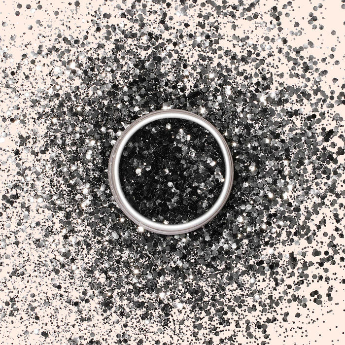 A silver ring rests on a surface sprinkled with Manucurist Biodegradable Glitter in black and silver, creating a sparkling, festive scene.