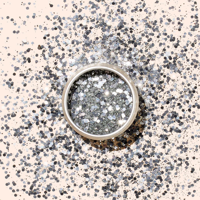 A round container of Manucurist Biodegradable Glitter rests on a pale surface, with extra glitter radiating around it—an eco-friendly and sparkling choice for creative nail art designs.
