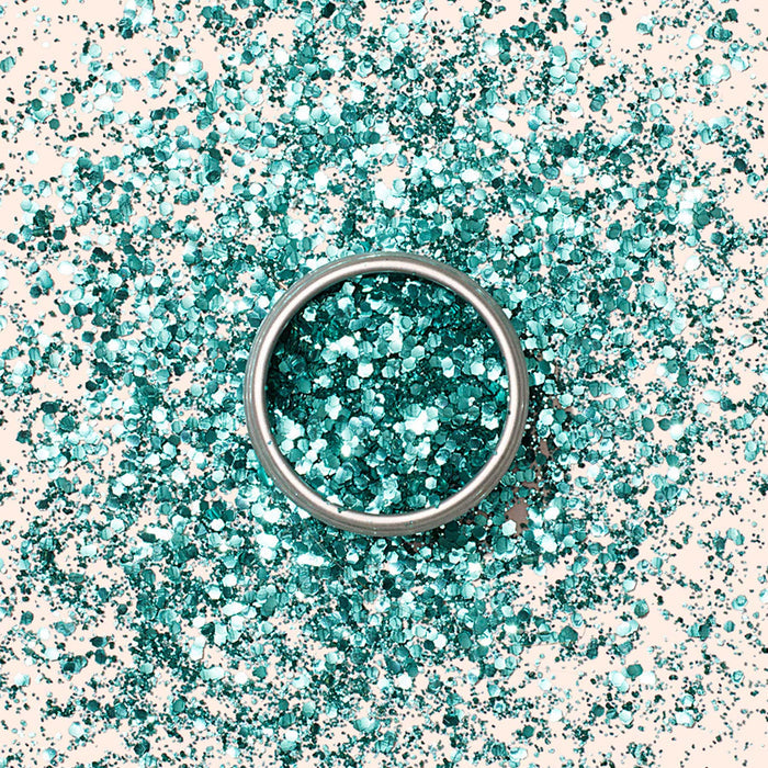 A ring sits atop a pile of Manucurist Biodegradable Glitter in turquoise, with scattered glitter on a light, neutral background.