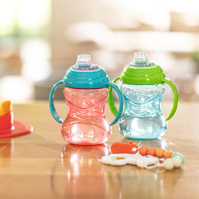 Two Nuby Clik-It Soft Spout Grip N' Sip Trainer Cups (Blue/Green, 2 Pack) sit on a wooden table. Each leak-proof cup has an easy-grip design and soft silicone spout, perfect for little hands. Baby toys and a blurred background complete the scene.