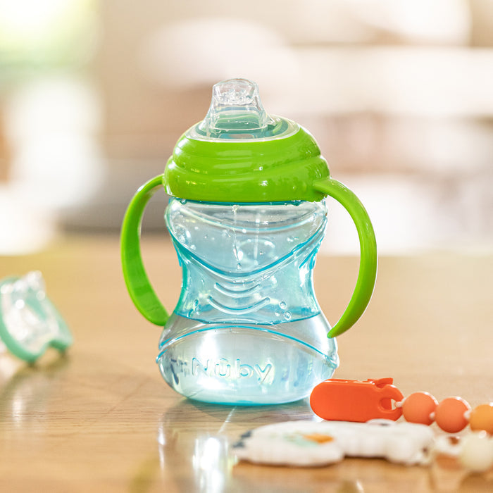 The Nuby Clik-It Soft Spout Grip N' Sip Trainer Cup (2 Pack) in blue/green with handles and a silicone spout rests on wood, accompanied by a pacifier and beaded clip, with a bright, softly blurred indoor background.
