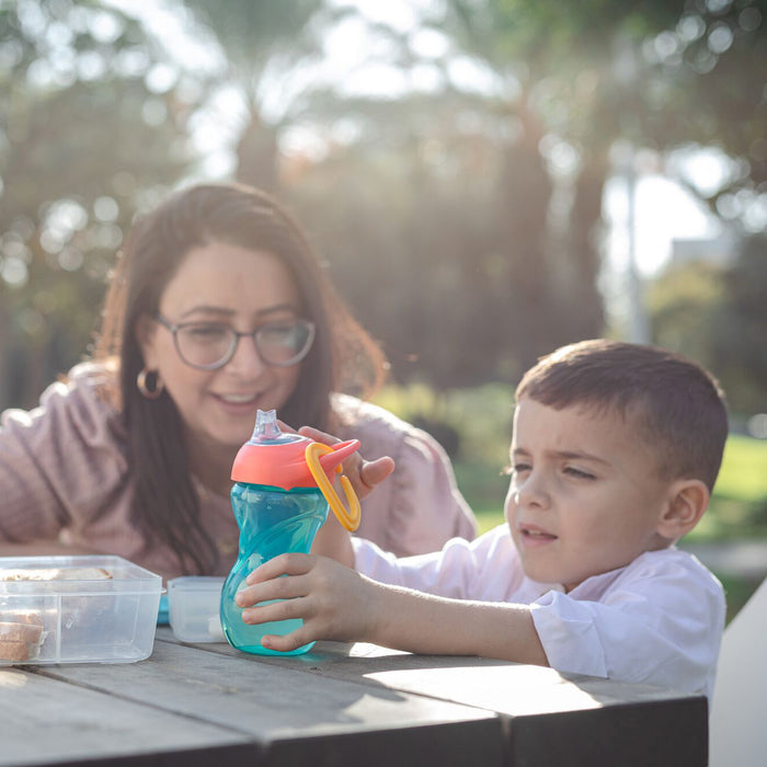 A woman and young boy sit outdoors. The boy reaches for a Clik-it Travel Sippy Cup with Carabiner (pink lid) as the woman smiles. Food containers are on the table, with trees in the background.