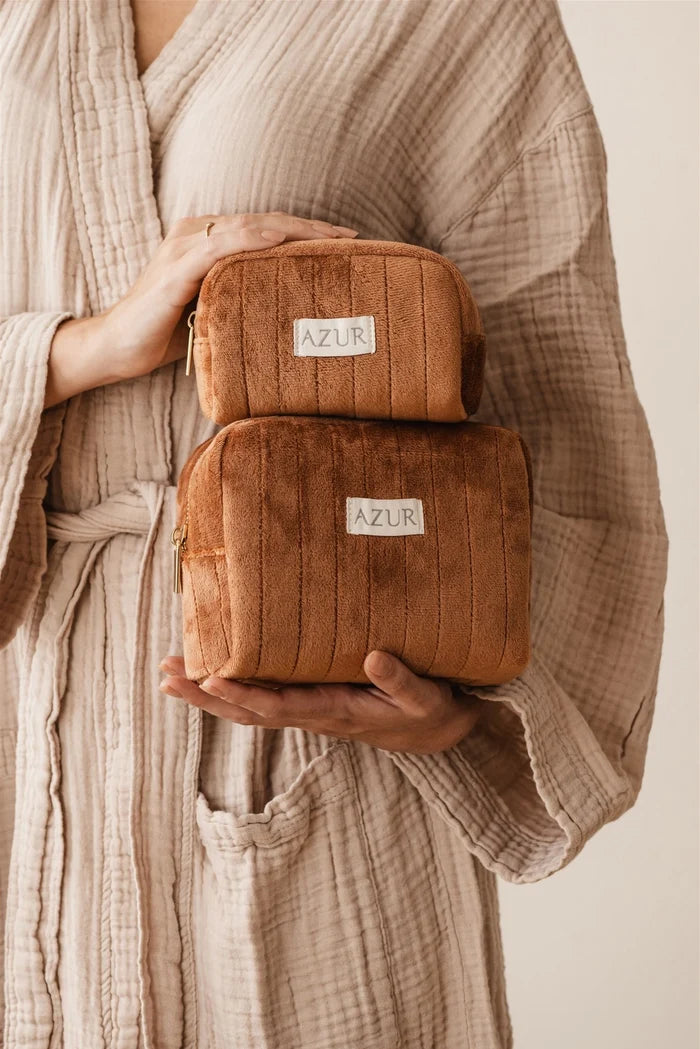Person holding two brown toiletry bags with 'AZUR' label, wearing a beige robe.