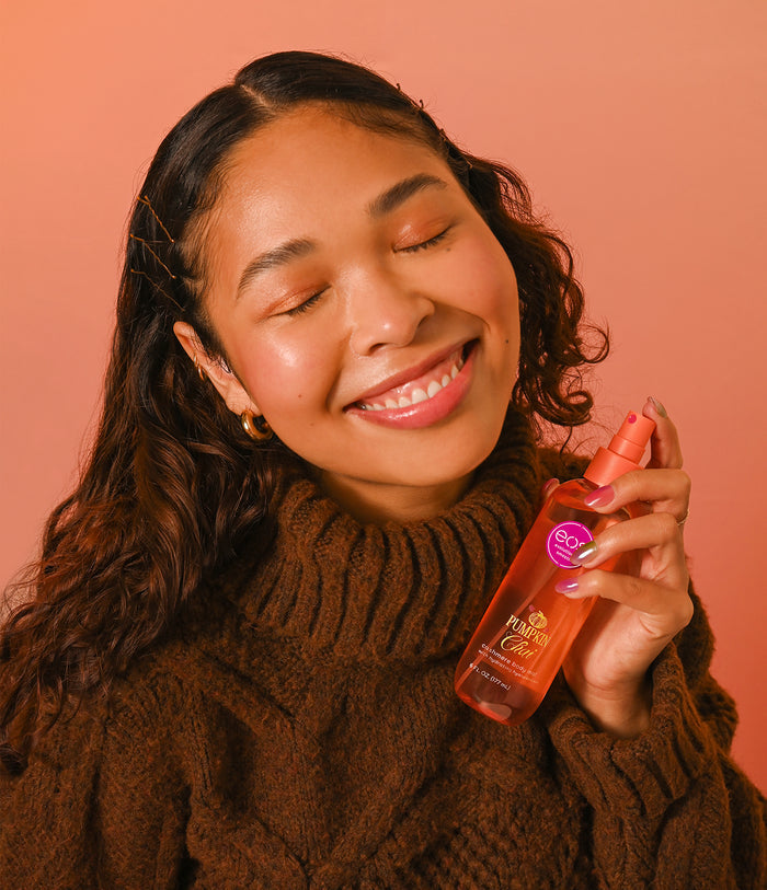 A woman with closed eyes and a smile holds a bottle of hair oil against a solid peach-pink background. She is wearing a brown cable-knit turtleneck sweater.