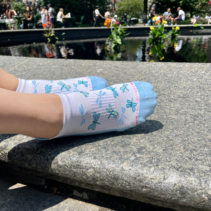 Ankle compression socks with a dragonfly design, displayed on a stone surface near a pond.