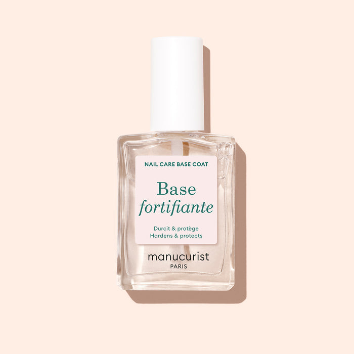 A clear glass bottle of Manucurist Force Nail Hardener with a white cap stands against a pale peach background. The label reads "Hardens  Protects" in French and English.