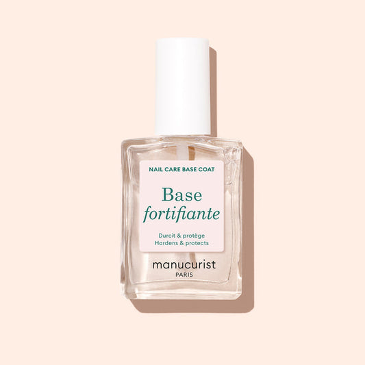 A clear glass bottle of Manucurist Force Nail Hardener with a white cap stands against a pale peach background. The label reads "Hardens  Protects" in French and English.