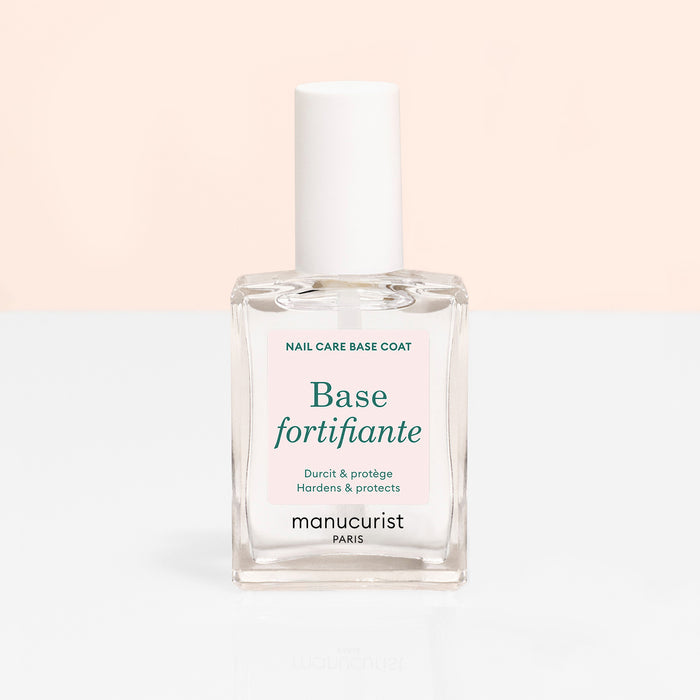 A clear glass bottle of Manucurist Force Nail Hardener with a white cap and label reading "Base fortifiante" and "Hardens  protects," set against a soft beige and white background.
