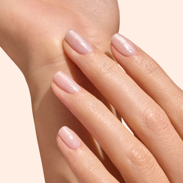 A close-up of a neatly manicured hand features shimmery pale pink nails finished with Manucurist's Glitter Top Coat, gently resting on the other wrist against a soft beige background.
