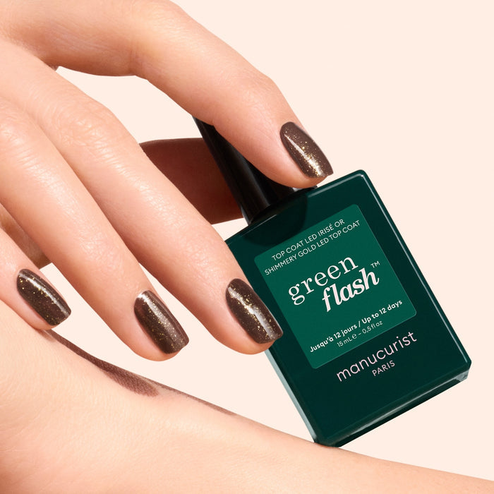 A hand with short, neat nails painted in shimmery brown with gold and silver shimmer holds a dark green bottle of Manucurist Glitter Top Coat against a light background.