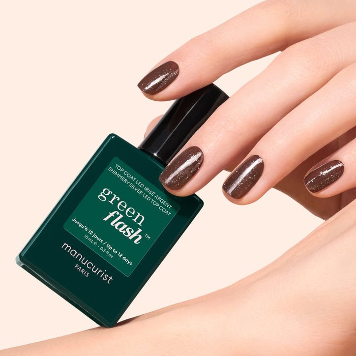 A hand with brown, glittery nail polish holds a green bottle of Glitter Top Coat by Manucurist against a light beige background, adding gold and silver shimmer for a long-lasting manicure.