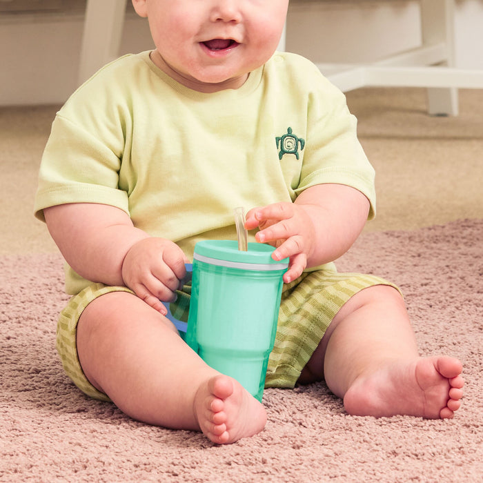 A baby in a yellow outfit sits on a pink carpet, holding the Insulated Tiny Tumbler | Seafoam with a silicone straw sippy cup. Only the baby's bottom half of the face and body are visible.