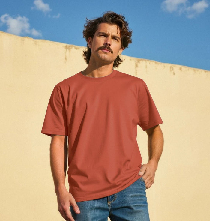 Man wearing a red t-shirt and blue jeans standing against a beige wall with a blue sky.