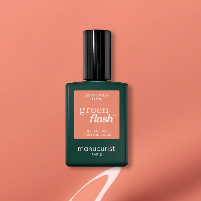 A bottle of Manucurist Green Flash gel polish in Peach rests on a peach background with a painted white swoosh underneath. The label highlights instant dry and extra long wear, making it an ideal summer tint.