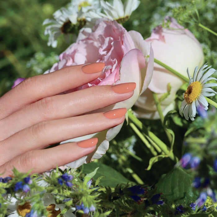 A hand with neatly manicured nails in Manucurist's Peach shade gently touches a light pink peony, surrounded by daisies and green foliage in a garden.