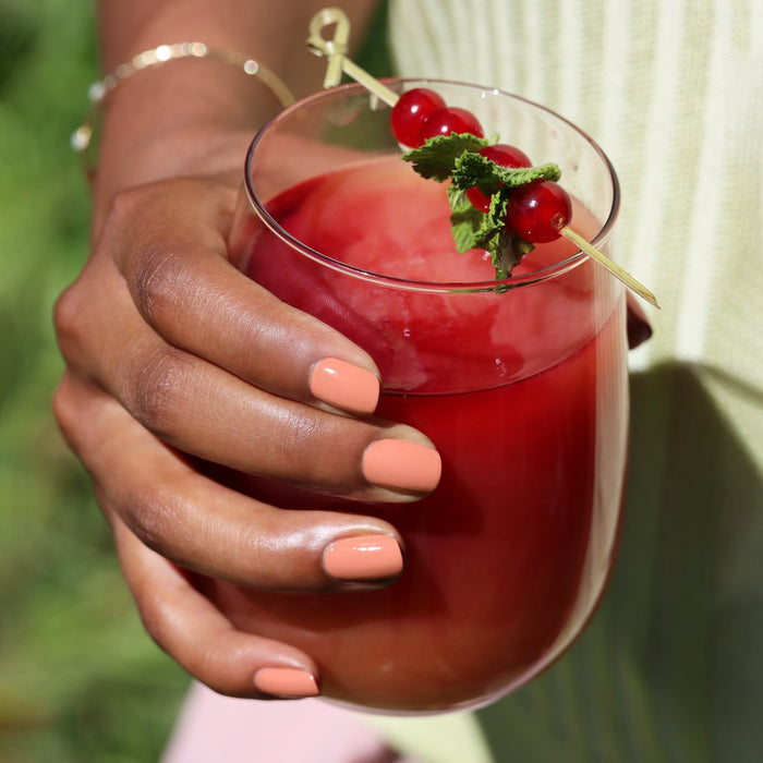 A person with Manucurist Peach nail polish holds a glass of red fruit juice topped with mint and red berries on a skewer. Blurred green foliage appears in the background.