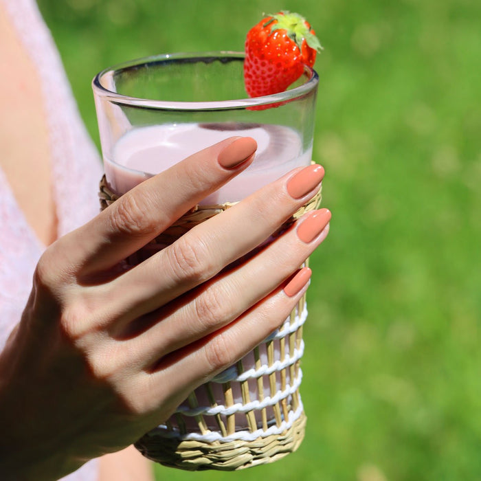 A hand with Manucurist Peach nails holds a glass of pink strawberry milk, garnished with a fresh strawberry and wrapped in a woven holder, set against a blurred green outdoor background.