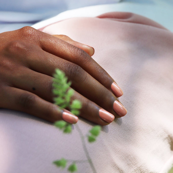 A close-up of a hand with neatly manicured Manucurist "Peach" nails rests on soft, pale pink fabric, with a small, blurred green fern in the foreground.