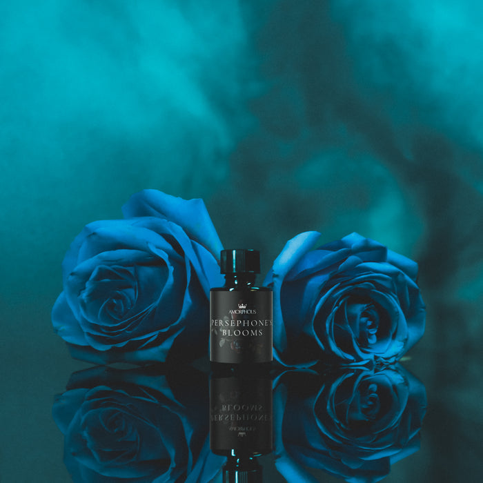 Persephone Blooms perfume bottle between two blue roses on a reflective surface with a teal background