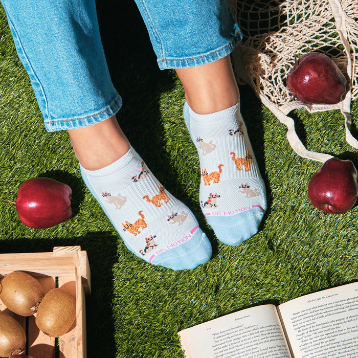Ankle compression socks with cat designs, worn by a person sitting on grass, surrounded by apples and a book.