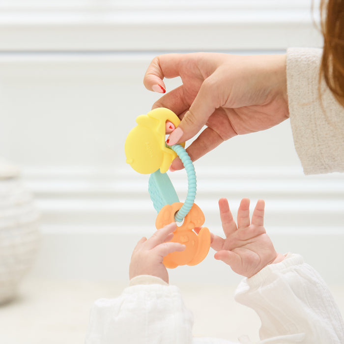 A woman’s hand holds the Silicone Bunny Basket Teether above a baby’s outstretched hands. The baby, dressed in white, reaches up toward the colorful bunny-shaped silicone teething toy.