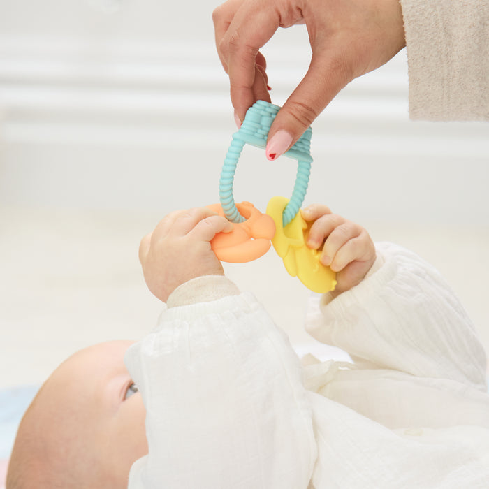 A baby in a white outfit reaches for the Silicone Bunny Basket Teether held by an adult, grasping the colorful toy with both hands as a soft, blurred background creates a gentle atmosphere.