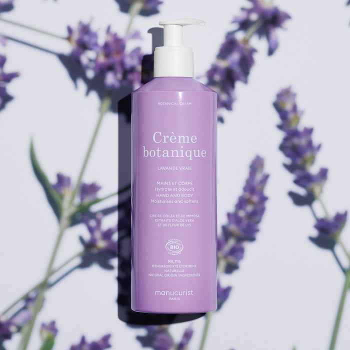 A purple bottle of Manucurist's Soothing Lavender Hand  Body Cream - Cosmos Organic with a white pump sits on a white surface, framed by lavender flowers and shadows. The French label highlights its botanical essence and hydrating formula.