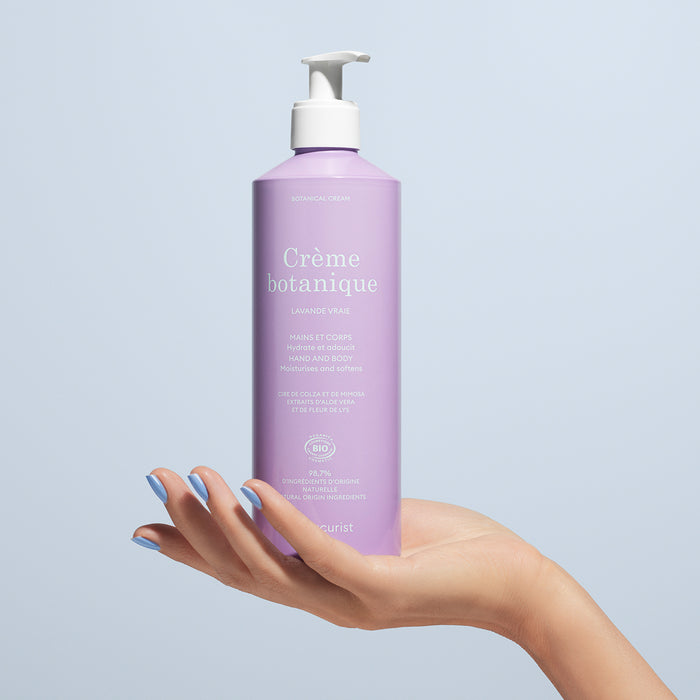 A hand with painted nails holds a lavender pump bottle labeled Soothing Lavender Hand  Body Cream - Cosmos Organic by Manucurist, an organic moisturizer for hands and body, set against a light blue background.