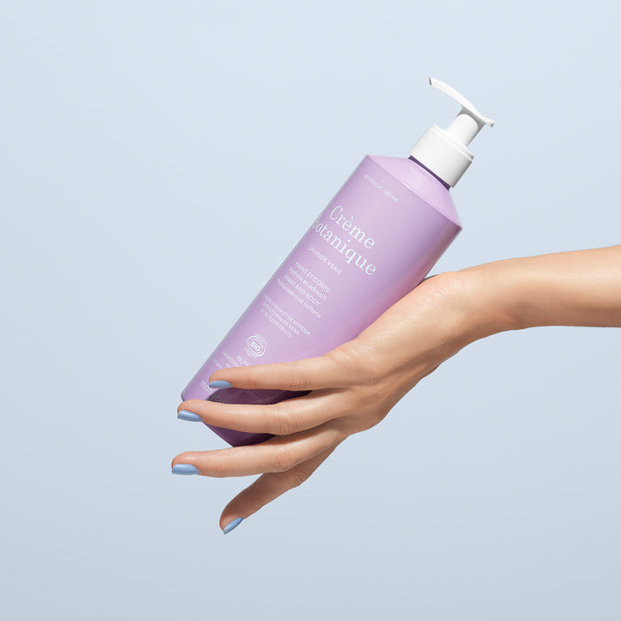 A hand with blue-painted nails holds a lavender-colored bottle of Manucurist Soothing Lavender Hand  Body Cream - Cosmos Organic with a white pump against a light blue background.