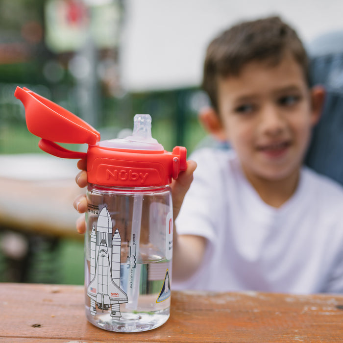 A young boy in a white shirt holds a clear water bottle with a red lid and straw. The bottle features a rocket and space-themed design, perfect for little astronauts, as he sits at a wooden table outdoors.