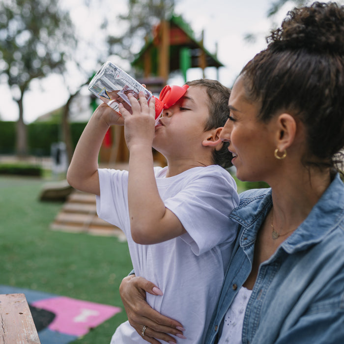 A young boy sips from a Nuby US Thirsty Kids NASA Flip-It Blast Cup while sitting on a woman's lap at a park. She smiles at him, and playground equipment appears in the background.