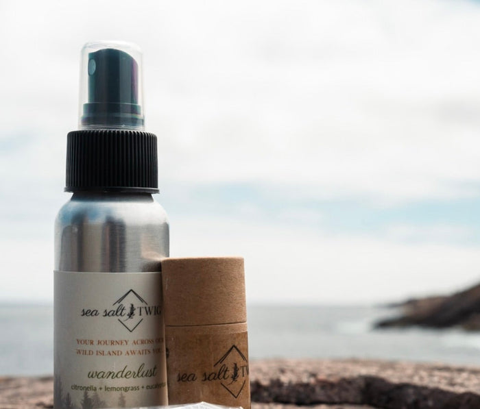 A set of skincare products including a body mist and body stick, displayed on a rock with a coastal background.