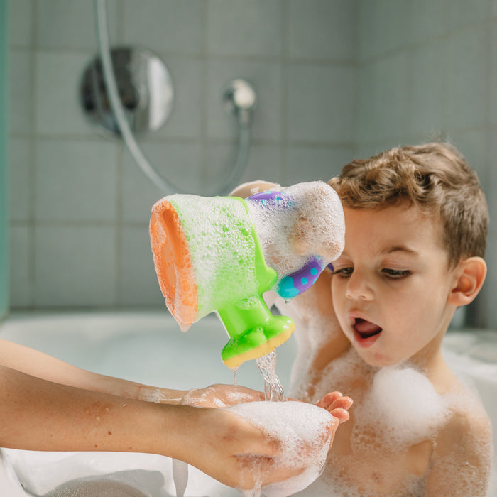 A young child in a bubble-filled bathtub pours water from the Watering Can Bath Toy onto an adult’s hand, both surrounded by bubbles and tiles.