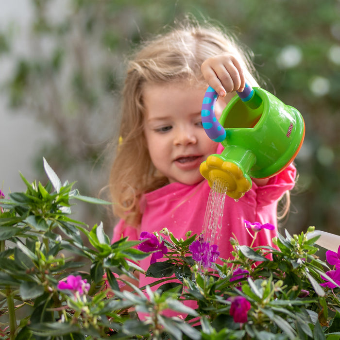 A young child with long blonde hair and a pink shirt waters blooming purple flowers using the Nuby Watering Can Bath Toy, surrounded by lush green leaves.